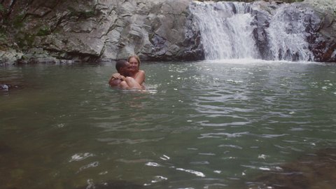 Young beautiful couple going swimming holding each other in secluded river near waterfall. Wide shot on 4k RED camera.