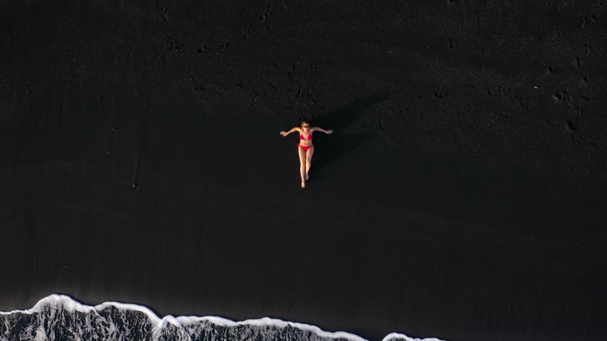 Top view of a woman in a red swimsuit lying on a black beach on the surf line. Coast of the island of Tenerife, Canary Islands, Spain. | Shutterstock HD Video #1040041826