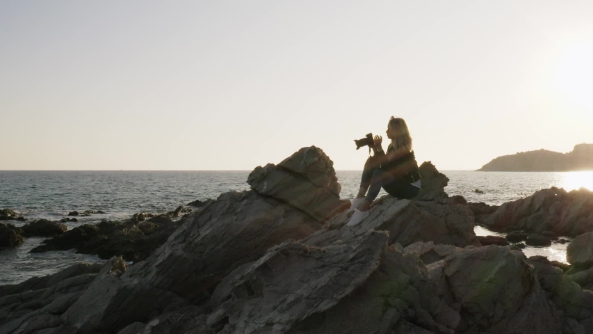 Slow motion of young grunge stylish woman photographer with DSLR camera taking pictures of the sea during sunset while sitting on the rocks.