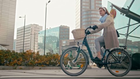 Millennial Business Woman Rides Bicycle in City Center in Modern Street in Autumn Day. Urban Cycle Chic and Ecological Transportation by Bike. 4K Slow Motion Long Tracking Shot with Copy Space