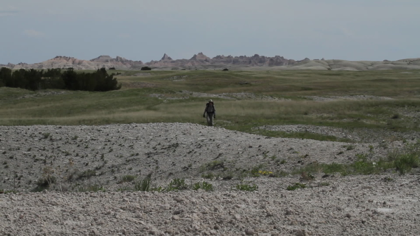 A male hiker walks across terrain and past camera in Badlands National Park, South Dakota