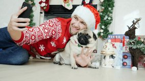 Man hug funny pug dog in christmas costume wears Santa hat and use mobile phone, calls friends on video connection, New Year and Christmas concept