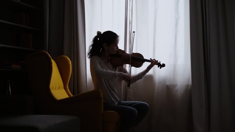 Beautiful girl musician plays on the background of a window at home, sitting on a soft chair. Shooting in slow motion.
