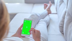 Hight quality 10bit footage of Young woman in white jeans uses SmartPhone with green screen laying on sofa at home. Made from 14bit RAW