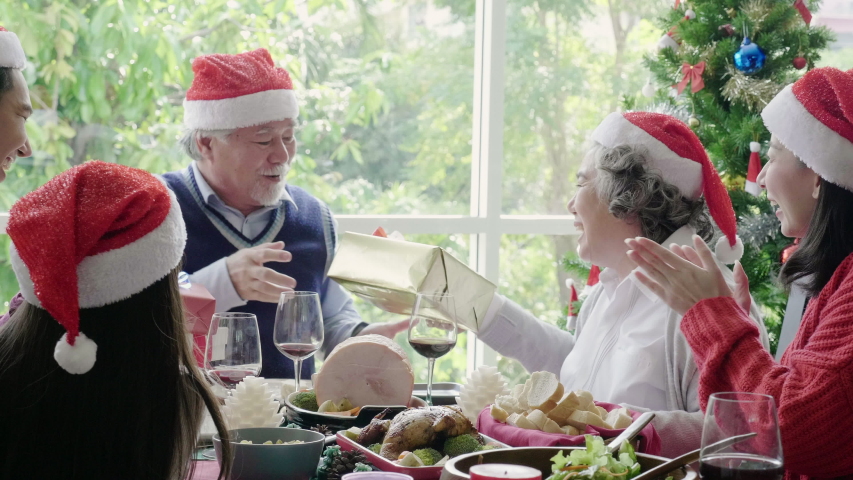 Asian family is eating a Christmas dinner with Grandfather, grandmother, parents and daughters sitting happily talking. A gift was given to Grandpa. People wear red Santa hats. New year celebration