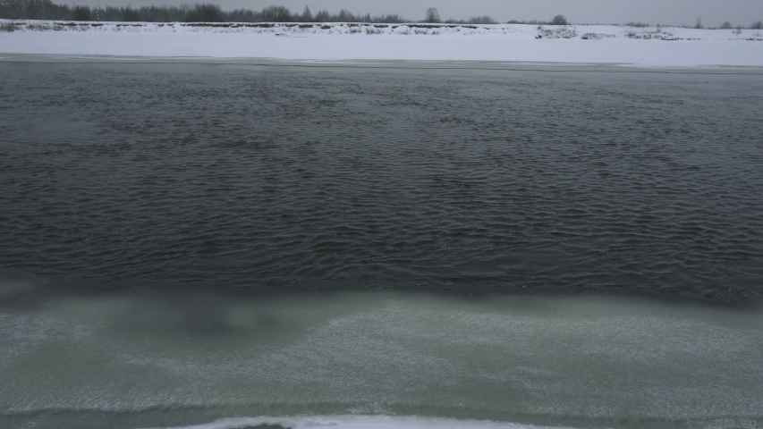 River flows in winter and snowing, ice along the edges of the bank in daytime. Nature background. Dangerous water for fishing