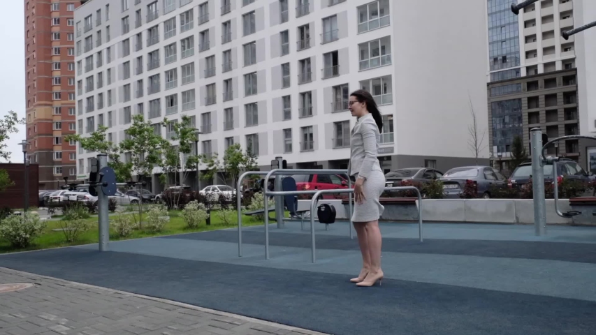 A woman in a business suit and heels does a sit-up outside. Business woman in glasses doing exercises during the break. The girl is engaged in fitness in the courtyard of the house on the playground.