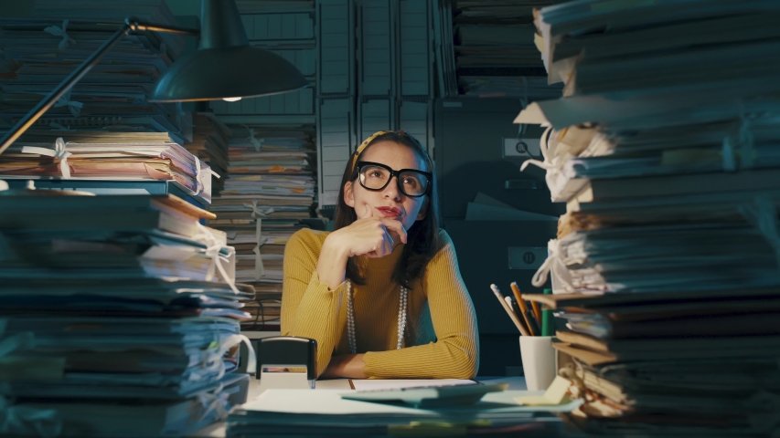 Young businesswoman sitting at office desk and thinking with hand on chin, she is surrounded by piles of paperwork Royalty-Free Stock Footage #1040075735