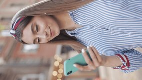 Verlical Video. Young Charming Caucasian Girl with Brown hair and Headband Wearing Blue and White Striped Dress Using her Smartphone Saying WoW and Smiling Standing at the City Street