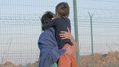 happy mother young woman and little poor child in dirty clothes refugees meeting on state border with fence hugging