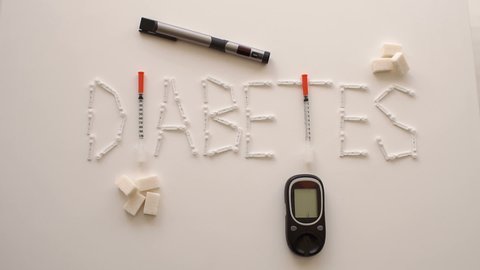 The word DIABETES is from lancets, insulin syringes, glucometer and insulin syringe pens on a white background, the child's hand puts a sugar cube next to the word. Slow motion. Theme of diabetes.