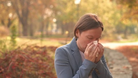Businesswoman nose-blowing paper tissue in autumnal park during the work break, handheld footage