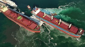 Aerial drone video of tug boats manoeuvring industrial bulk carrier by pushing or pulling in Mediterranean shipyard