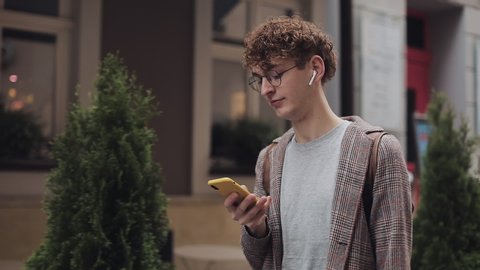 Young Hipster Guy in Glasses with Earphones Wearing Checked Jacket Having Backpack. Chatting with Friends, Using his Smartphone. Walking at Old City Background. Student, Tourist Concept.