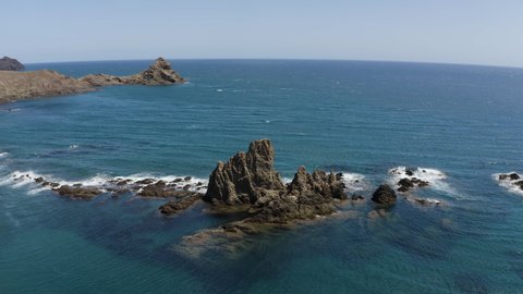 Aerial drone footage of blue sea with little waves and rocks at beautiful Cabo de Gata in Andalusia, during day. Blue sky, Almeria, Spain, bluish color, 4K UHD. Drone turning around rock.