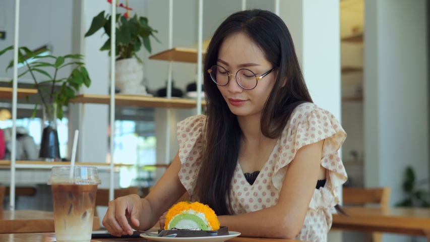 Young asian woman is biting off piece of dessert, drinking coffee and looking at a camera. Portrait Thai woman eating cake  in cafe close up.