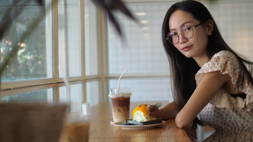 Young asian woman is biting off piece of dessert, drinking coffee and looking at a camera. Portrait Thai woman eating cake  in cafe close up.