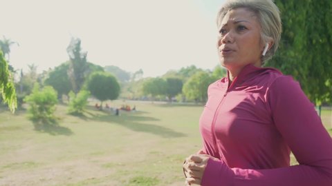 slow motion gimbal tracking shot on attractive middle aged lady running happy at city park . beautiful and sporty 40s woman exercising doing jogging workout enjoying healthy lifestyle and fitness
