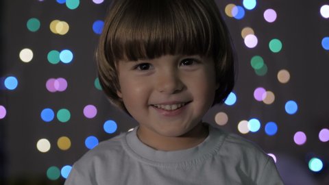 Cute Little Boy With Standing Against Glowing Christmas Lights Smiling at Camera. Portrait Child Looking at Camera. Christmas New Year Background. Lovely Boy Posing On Christmas Lights Background