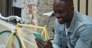Cheerful African American Guy Having a Video Call via his Smartphone, Waving, Smiling while Sitting near Stylish Bike with Old Metal Fance at the Background. Side View. Close Up