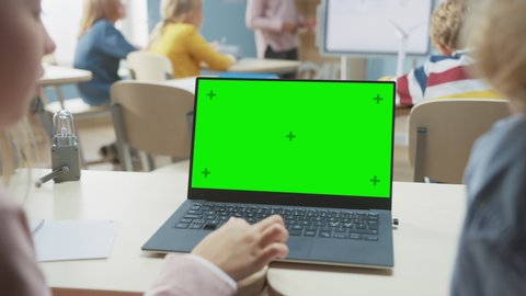 Elementary School Classroom: Over the Shoulder View of a Two Kids Using Green Mock-up Screen Laptop in Class. In the Background Teacher Explains Lesson to Diverse Class