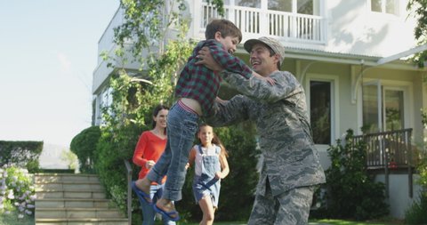 Rear view of a young adult mixed race male soldier in the garden outside his home, kneeling and picking up his young son, who runs to him holding a US flag, with his mother and sister holding hands