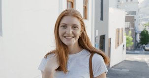 Portrait close up of a young Caucasian woman with long red hair standing in a sunny city street smiling to camera, slow motion 