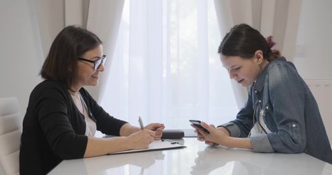 Teenage patient talking with female professional psychotherapist, sitting at a table in the office. Girl talking, looking at a smartphone and laughing