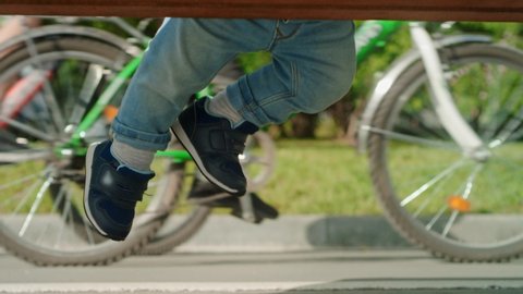 Close-up: legs of a small child in shoes sitting on a Park bench on a Sunny summer day. The kid cheerfully dangles his legs hanging over the ground. Family, children. Front view, Slow motion, 4K, RED