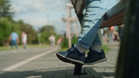 Close-up: legs of a small child in shoes sitting on a Park bench on a summer day. The kid cheerfully dangles his legs hanging over the ground. Family, children. Side view, Slow motion, 4K, RED