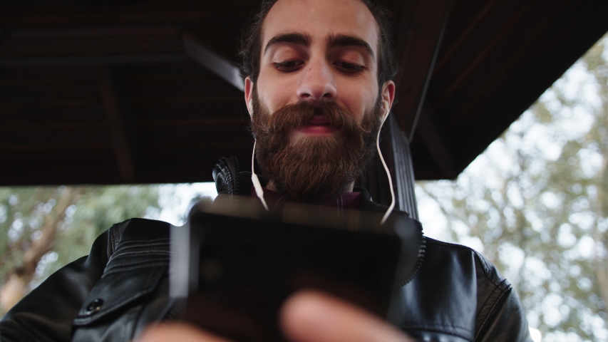 Young handsome bearded man is texting,listening music on phone with earphones and smiling. 
He is sitting in park outdoors. | Shutterstock HD Video #1040172749