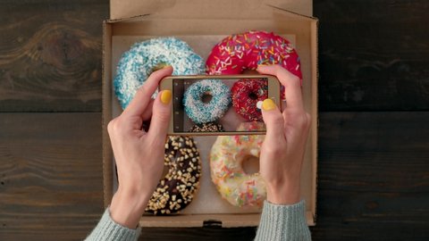 Top view - Woman's hands take pictures with smartphone of delicious fresh donuts in box on wooden table. 4k. Concept of food photo blogging.