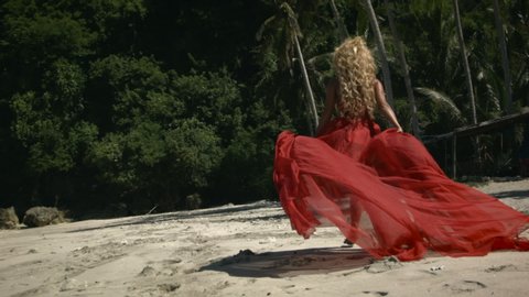 Young happy woman with curly blonde hair in elegant fluttering long red dress walking and jumping on beach. Trees, umbrellas and palms on background.
