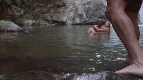 Man dives into lagoon while young beautiful coupleswim holding each other in secluded river near waterfall. Wide shot on 4k RED camera.