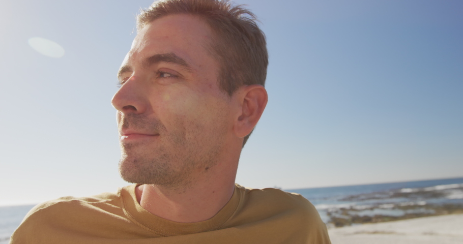 Front view close up of a happy young Caucasian man sitting on sunny beach looking around and smiling, slow motion