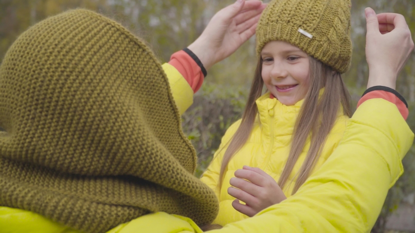 Adult Caucasian woman fixing mustard hat on little girl's head. Mother and daughter dressed in yellow coats laughing together. Family having fun outdoors