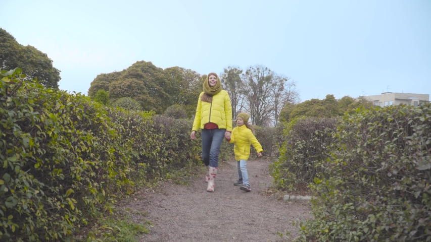 Happy Caucasian girl jumping along green bushes with her cheerful mother. Smiling mother resting with her little daughter outdoors. Cheerful family approaching the camera.