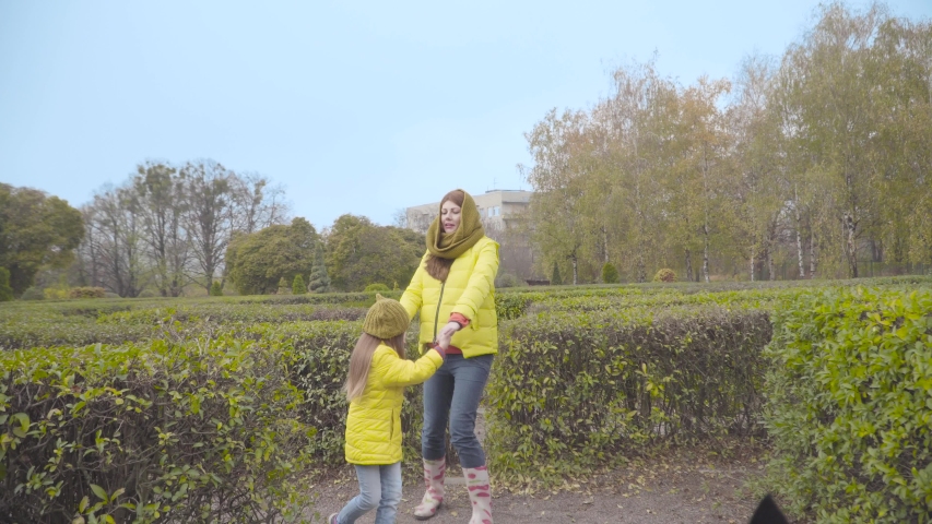Happy Caucasian family in yellow coats playing outdoors. Daughter spinning aroung her mother in the autumn park. Cheerful mom resting with her child.