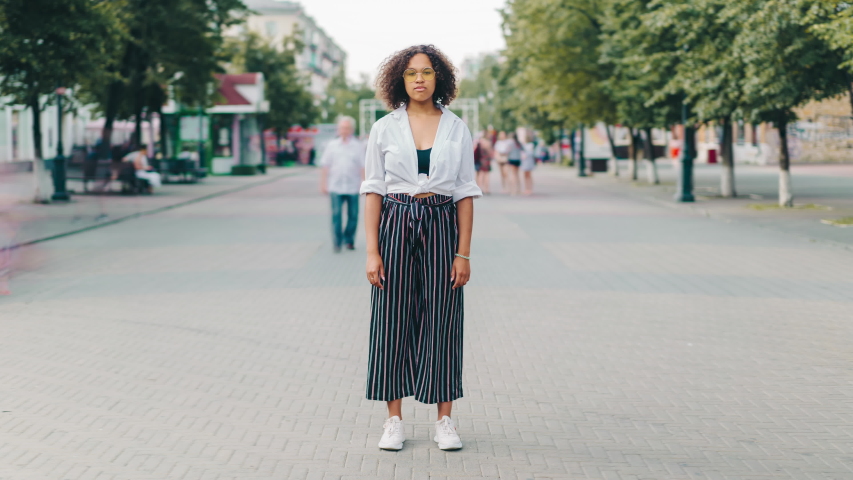 Zoom in time lapse of serious young Afro-American woman in glasses standing outdoors in city looking at camera. Modern youth and casual life concept.