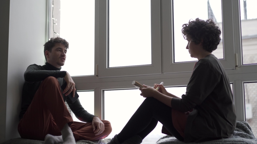 A young couple sits talking and chatting at a large window next to a Christmas tree in their cozy home. A girl draws a man or makes sketches.
