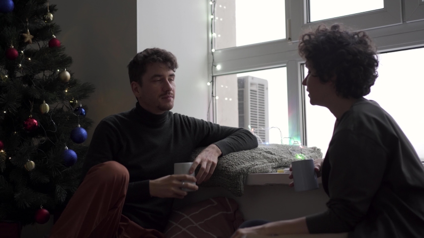 A young couple sits talking and chatting at a large window next to a Christmas tree in their cozy home. A man and a girl with hot drinks in mugs.
