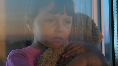 Sad embracing by the window. Child with tears on her face embrace a teddy bear near the window.