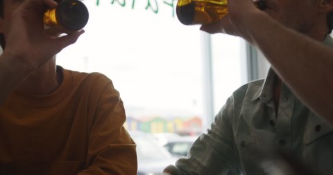 Front view mid section of a caucasian father and his young adult son sitting at a table in a cafe drinking bottles of beer and talking, the father with his hand on the shoulder of his son, slow motion