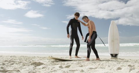 Side view of a caucasian father and his young adult son on a beach wearing wetsuits, the son standing on a surfboard and the father giving him some advice before surfing, slow motion