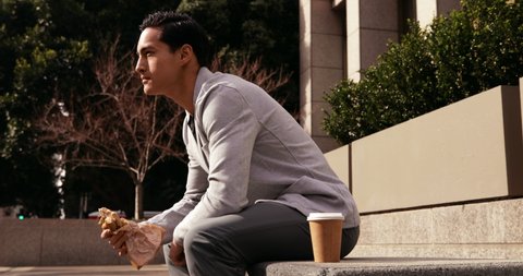 Side view of a young mixed race man sitting on a bench, eating sandwich with takeaway coffee next to him during a lunch break in the city street with building in the background