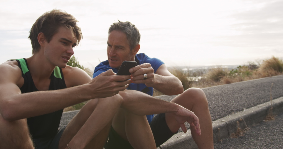 Side view close up of a caucasian father and his young adult son running together by the coast taking a break, sitting at the roadside looking at a smartphone and talking, slow motion