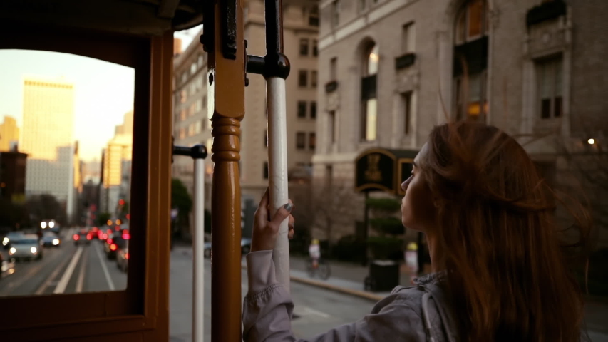 Rear view close up of a young Caucasian woman with long dark hair standing on the outside of a tram hanging on to a handle while travelling down a city street at dusk, slow motion | Shutterstock HD Video #1040192099