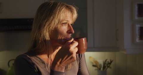 Caucasian woman drinking a cup of coffee at home, standing in her kitchen, looking straight ahead and smiling. Social distancing and self isolation in quarantine lockdown for Coronavirus Covid19