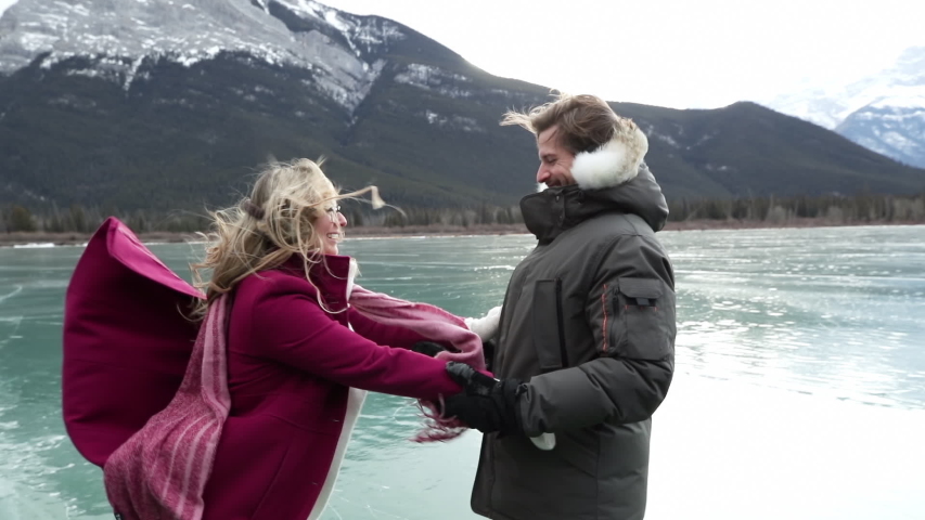Side view close up of a millennial Caucasian couple ice skating on a frozen lake, holding hands, the woman turning on the ice, and both smiling at each other, with mountains in the background