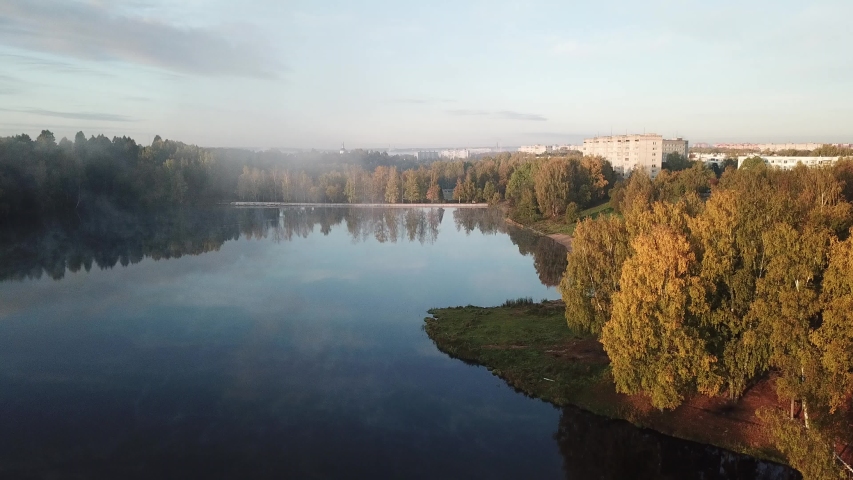 4K aerial summer early morning high quality video footage of pristine lush green pine tree surrounded forest lake with dark calm water reflecting clouds, a dam, island and a sand beach near small town